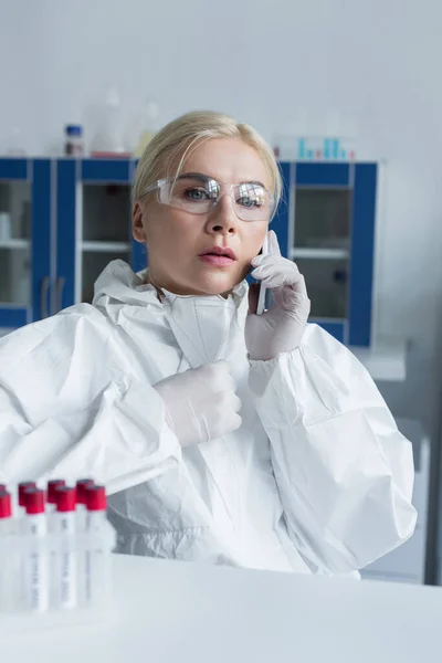 Scientist in protective suit talking on smartphone near blurred test tubes in lab — Fotografia de Stock