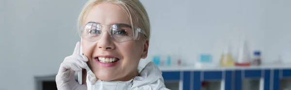 Cheerful scientist in protective goggles talking on cellphone in lab, banner — Stockfoto