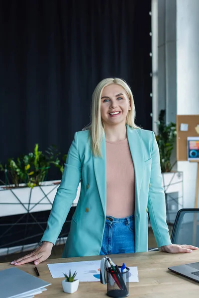 Smiling psychologist looking at camera near papers and laptop in consultation room - foto de stock