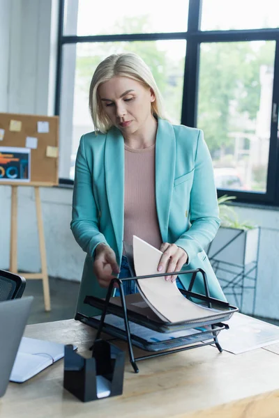Blonde psychologist holding paper folder near stationery and laptop in consulting room — Stock Photo