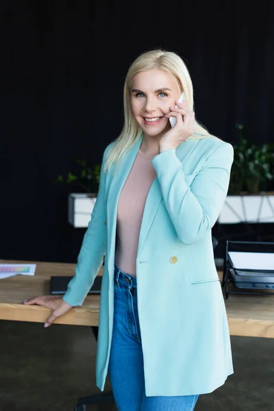 Cheerful psychologist talking on smartphone near table in consultation room — стоковое фото