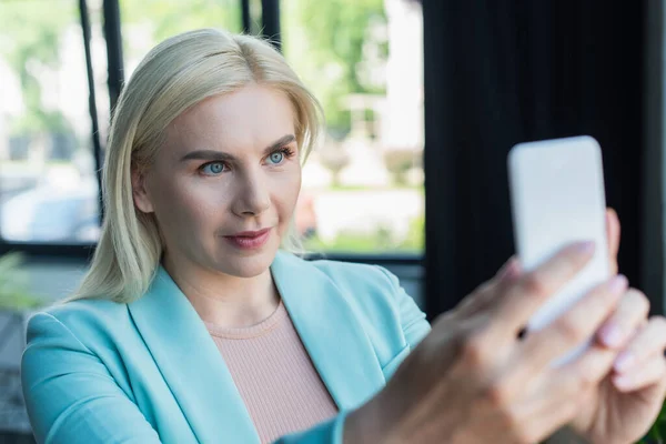 Blonde psychologist having video call on blurred smartphone in consultation room — стоковое фото