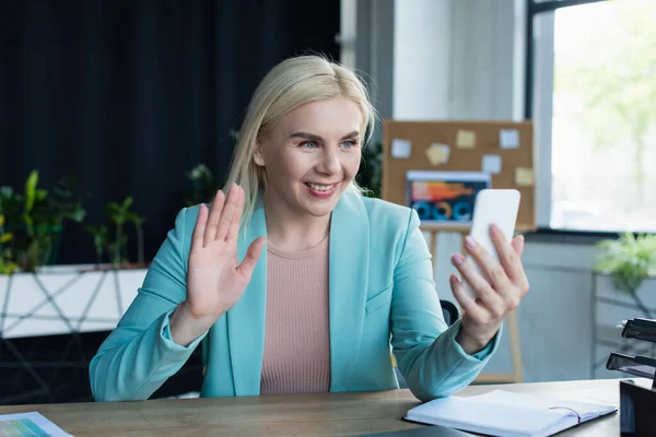 Smiling psychologist having video call on smartphone near notebook in consultation room — Foto stock