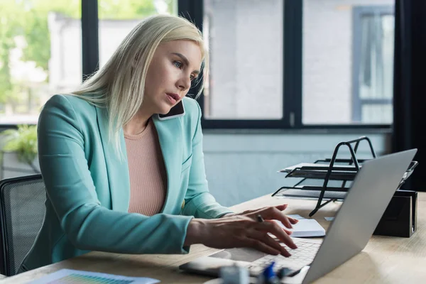 Blonde psychologist talking on smartphone and using laptop in consultation room — Fotografia de Stock