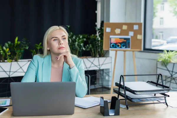 Pensive psychologist looking away near laptop and papers in consultation room — Photo de stock