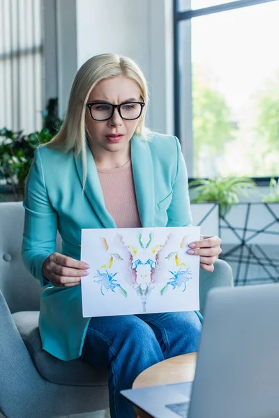 Focused psychologist holding Rorschach test during video call on laptop in consultation room — Fotografia de Stock