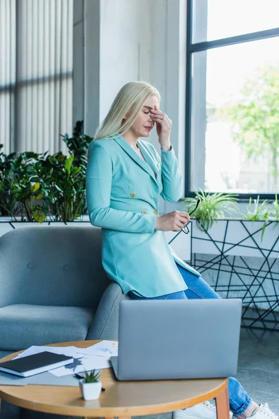 Tired psychologist holding eyeglasses near laptop in consultation room — Stock Photo