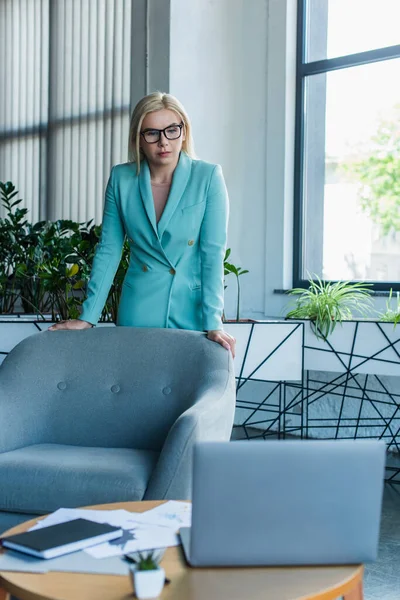 Psychologist in eyeglasses looking at laptop near armchair in consultation room — Foto stock