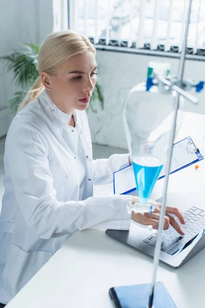 Scientist using laptop and holding clipboard near flask in lab — Stock Photo