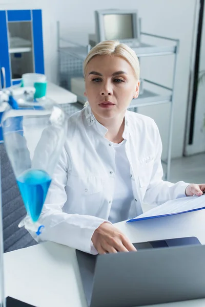 Scientist holding clipboard and looking at flask near laptop in lab — Fotografia de Stock