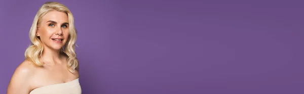 Happy blonde woman with bare shoulders looking at camera isolated on purple, banner — Stock Photo