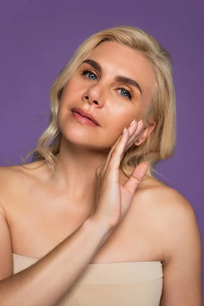 Blonde woman with bare shoulders touching cheek and looking at camera isolated on purple — Stock Photo