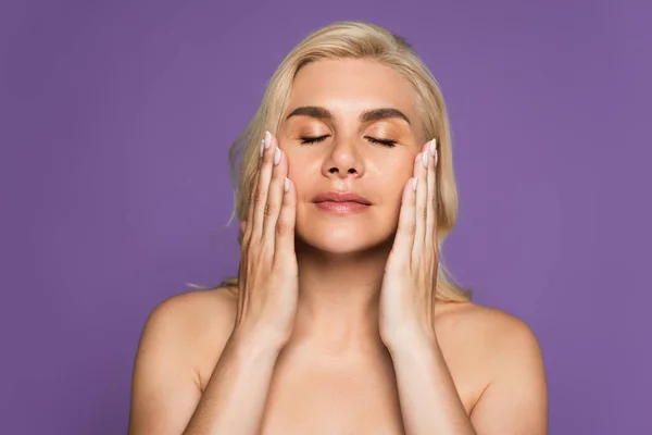 Blonde woman with bare shoulders and closed eyes applying cream isolated on purple — Stockfoto