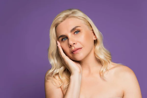 Sensual blonde woman with bare shoulders looking at camera while touching cheek isolated on purple — Foto stock