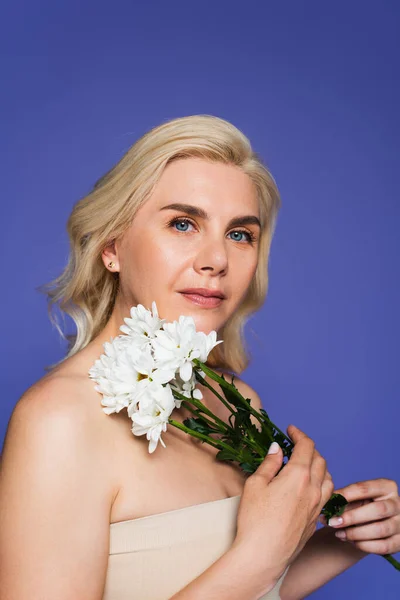 Blonde Frau mit blauen Augen, die in die Kamera schaut, während sie weiße Blumen isoliert auf Veilchen hält — Stockfoto