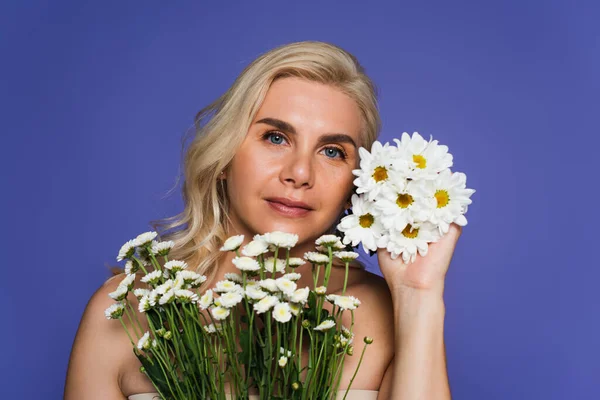 Блондинка з голими плечима тримає білі квіти і дивиться на камеру ізольовано на фіолетовому — стокове фото