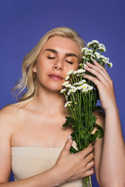 Donna sensuale e bionda con gli occhi chiusi che tiene fiori isolati su viola — Foto stock