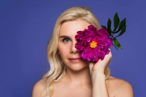 Blonde woman with bare shoulders covering eye with blooming flower and looking at camera isolated on purple — Foto stock