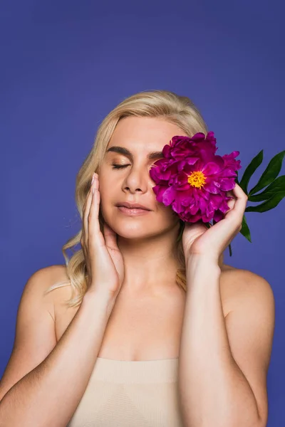 Blonde woman with bare shoulders covering eye with blooming flower isolated on purple — Stock Photo