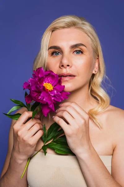 Mulher loira com olhos azuis segurando flor florescendo e olhando para câmera isolada em roxo — Fotografia de Stock