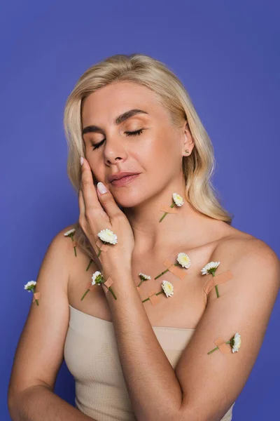 Sensual mujer rubia con los ojos cerrados y flores en flor en el cuerpo aislado en púrpura - foto de stock