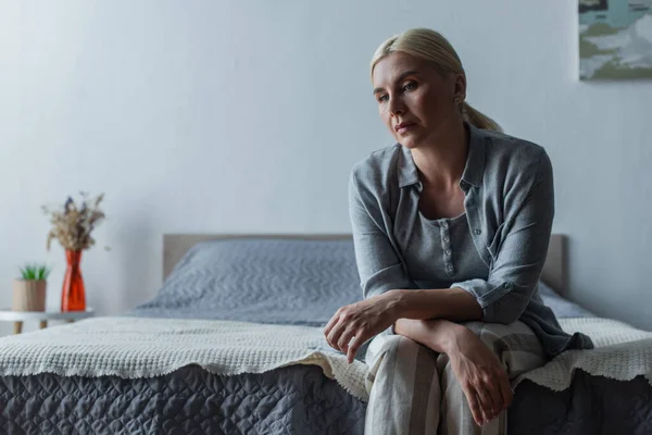Расстроенная блондинка с менопаузой сидит на кровати дома — стоковое фото