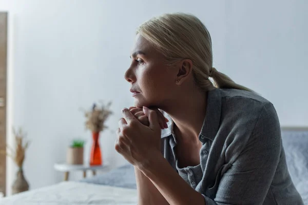 Side view of worried blonde woman with menopause looking away in bedroom - foto de stock