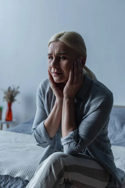 Депресивна блондинка з менопаузою торкається обличчя, сидячи на ліжку — стокове фото
