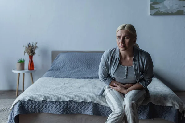 Upset blonde woman with menopause suffering from stomach ache in bedroom — Foto stock