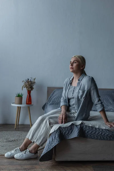 Full length of upset blonde woman with menopause looking away while sitting on bed — Photo de stock