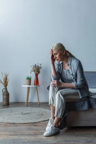 Volle Länge der müde blonde Frau mit Wechseljahren, die unter Kopfschmerzen leidet, während sie auf dem Bett sitzt — Stockfoto