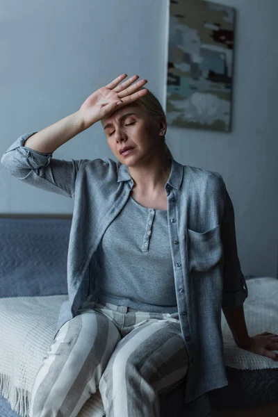 Mujer agotada con menopausia que sufre de dolor de cabeza mientras está sentada en el dormitorio - foto de stock