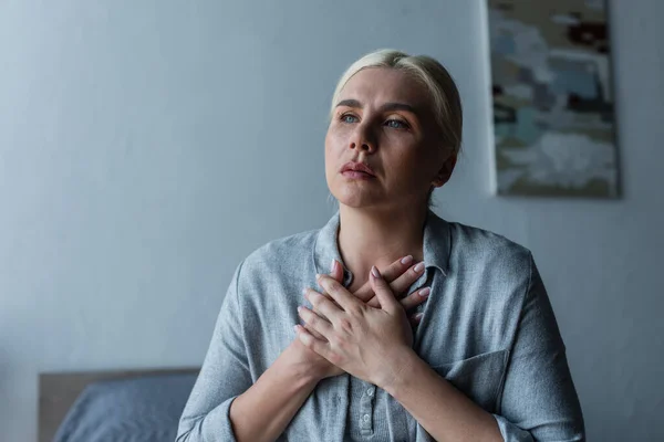 Mujer rubia con menopausia tocando el pecho mientras sufre de calor en el dormitorio - foto de stock