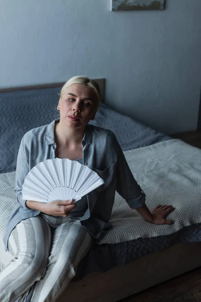 Mujer rubia con menopausia que sufre de calor y refrigeración con ventilador en el dormitorio - foto de stock