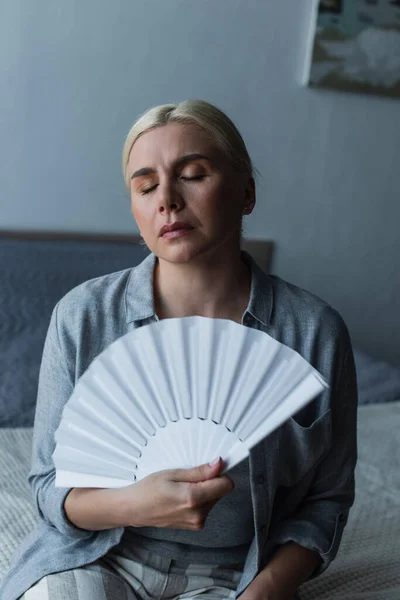 Displeased blonde woman with menopause suffering from heat and cooling with fan in bedroom — Stock Photo