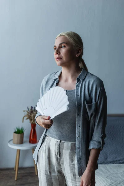 Blonde woman with menopause suffering from heat and holding fan in bedroom — Stock Photo