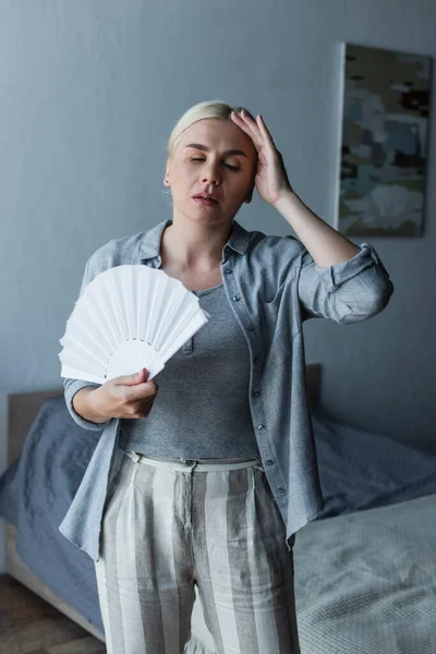 Exhausted woman with menopause suffering from heat and holding fan in bedroom — Stock Photo