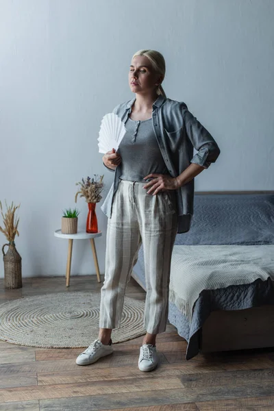 Full length of blonde woman with menopause suffering from heat and holding fan in bedroom — Fotografia de Stock