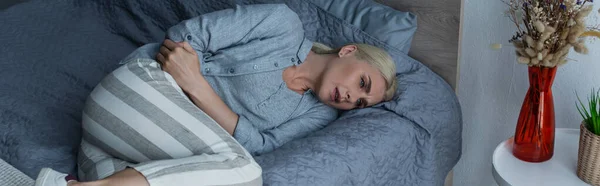 Blonde Frau mit Wechseljahren, die unter Bauchschmerzen leidet und im Bett liegt, Banner — Stockfoto