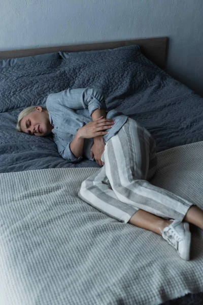 Donna bionda con menopausa che soffre di dolore addominale e sdraiata sul letto — Foto stock