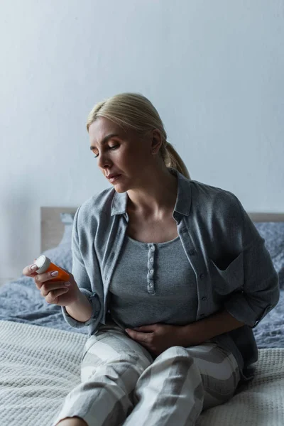 Femme blonde déprimée avec ménopause tenant bouteille avec des pilules et assise sur le lit — Photo de stock