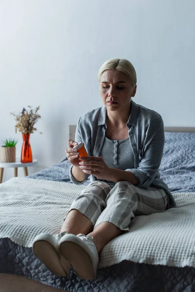 Displeased blonde woman with menopause holding bottle with pills and sitting on bed — Stock Photo