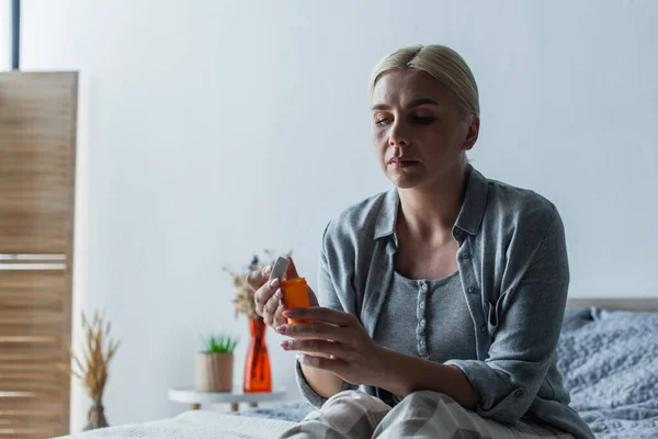 Mujer rubia cansada con la menopausia sosteniendo la botella con pastillas y sentado en la cama - foto de stock