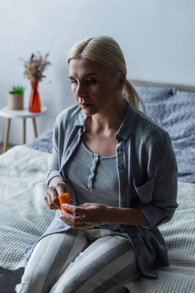 Mulher chateada com menopausa segurando frasco com medicação e sentado na cama — Fotografia de Stock