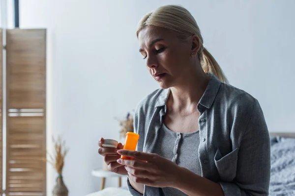 Mulher loira frustrada com menopausa segurando garrafa com pílulas e sentado no quarto — Fotografia de Stock