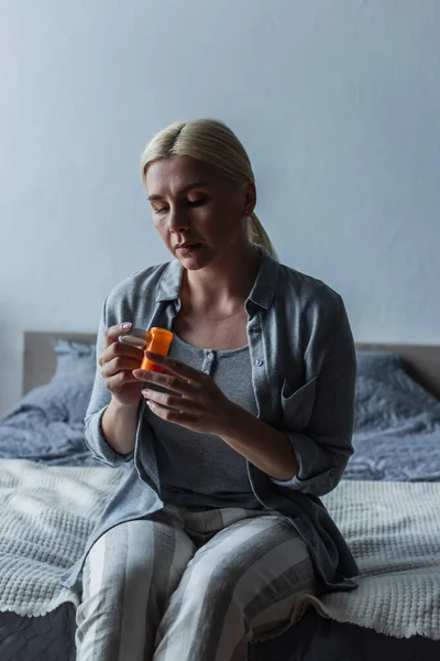 Сумна блондинка з менопаузою тримає пляшку з таблетками і сидить на ліжку — стокове фото