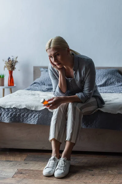 Upset blonde woman with menopause holding bottle with pills and sitting on bed — Stock Photo