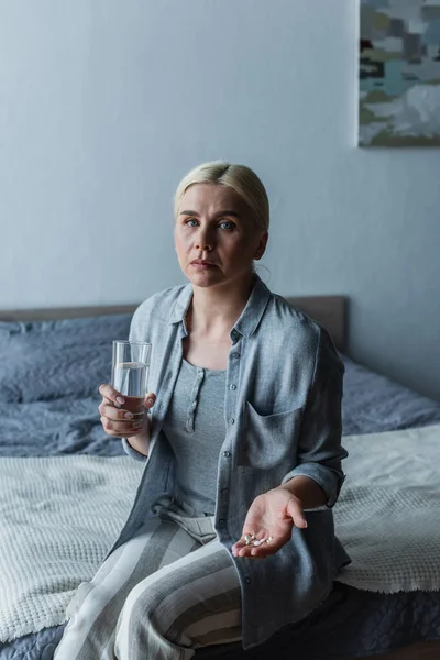 Жінка з менопаузою тримає таблетки і склянку води, сидячи на ліжку — стокове фото