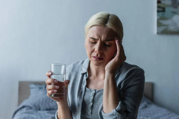 Blondine mit Wechseljahren leidet unter Kopfschmerzen und hält Glas Wasser in der Hand — Stockfoto