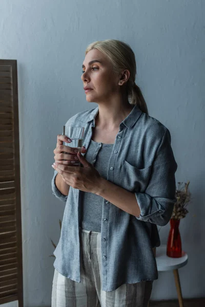 Mulher loira com menopausa segurando vidro de água e olhando para longe — Fotografia de Stock